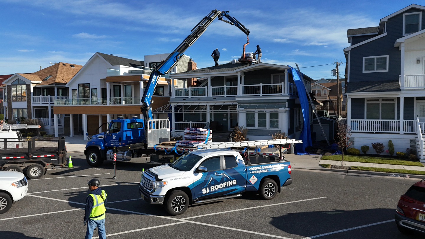 A truck with a crane delivers roofing materials to workers on a house roof, while another branded pickup and a worker stand in a residential neighborhood.
